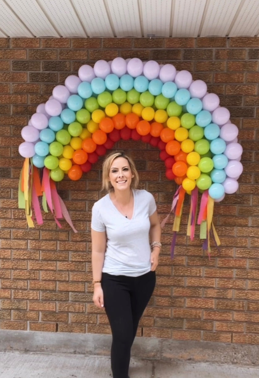 Young woman standing in front of a pastel coloured rainbow-shaped balloon arch smiling